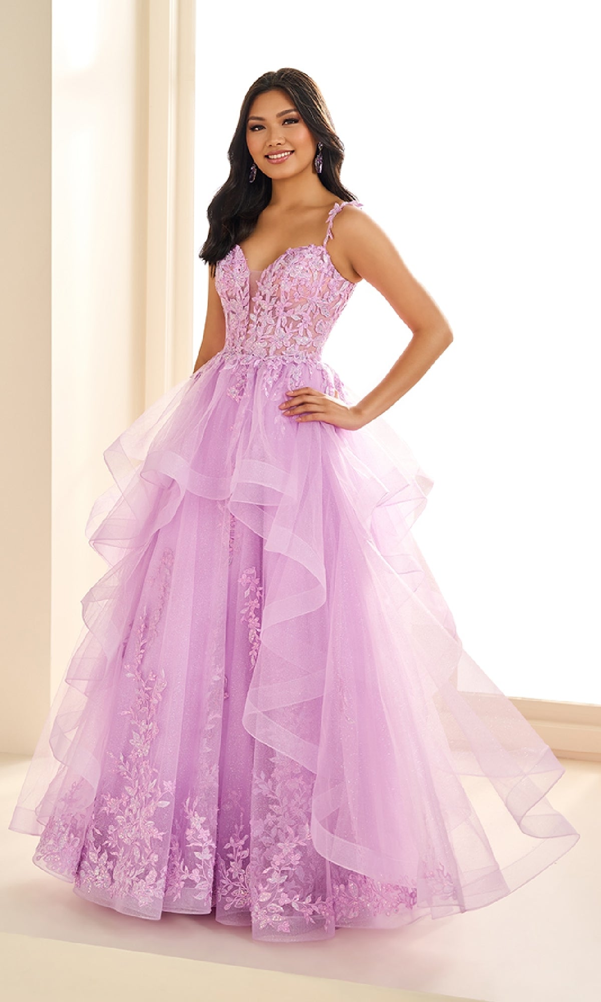 A woman stands indoors wearing the Ellie Wilde EW36081, a long lilac prom dress featuring floral lace appliqué, a fitted bodice with spaghetti straps, and a layered glitter tulle skirt.